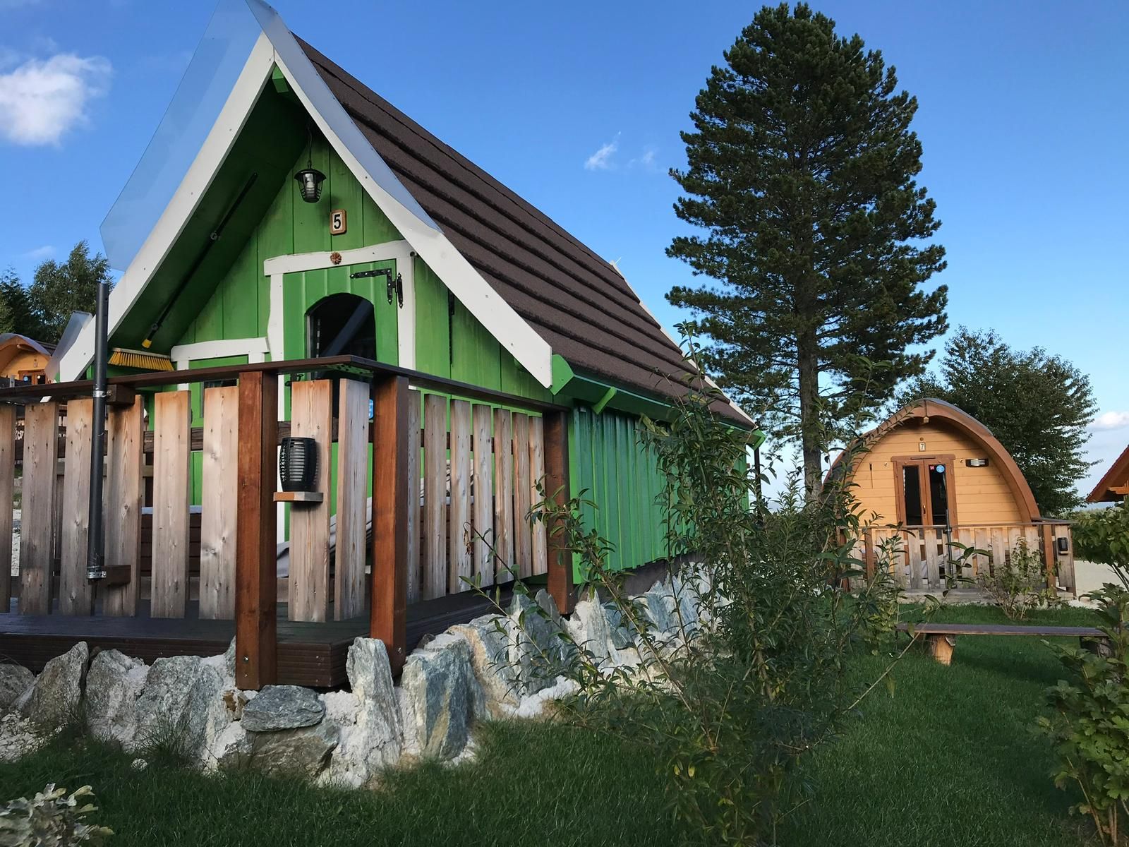 Two small wooden cabins in glamping style with a green and brown façade, surrounded by trees and grass.