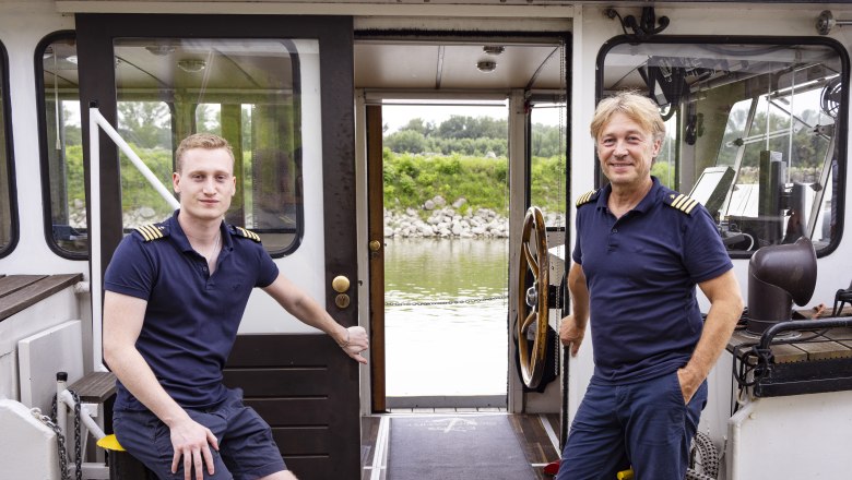 Two men in naval uniform on a ship, with a view of a river and green shore landscape.