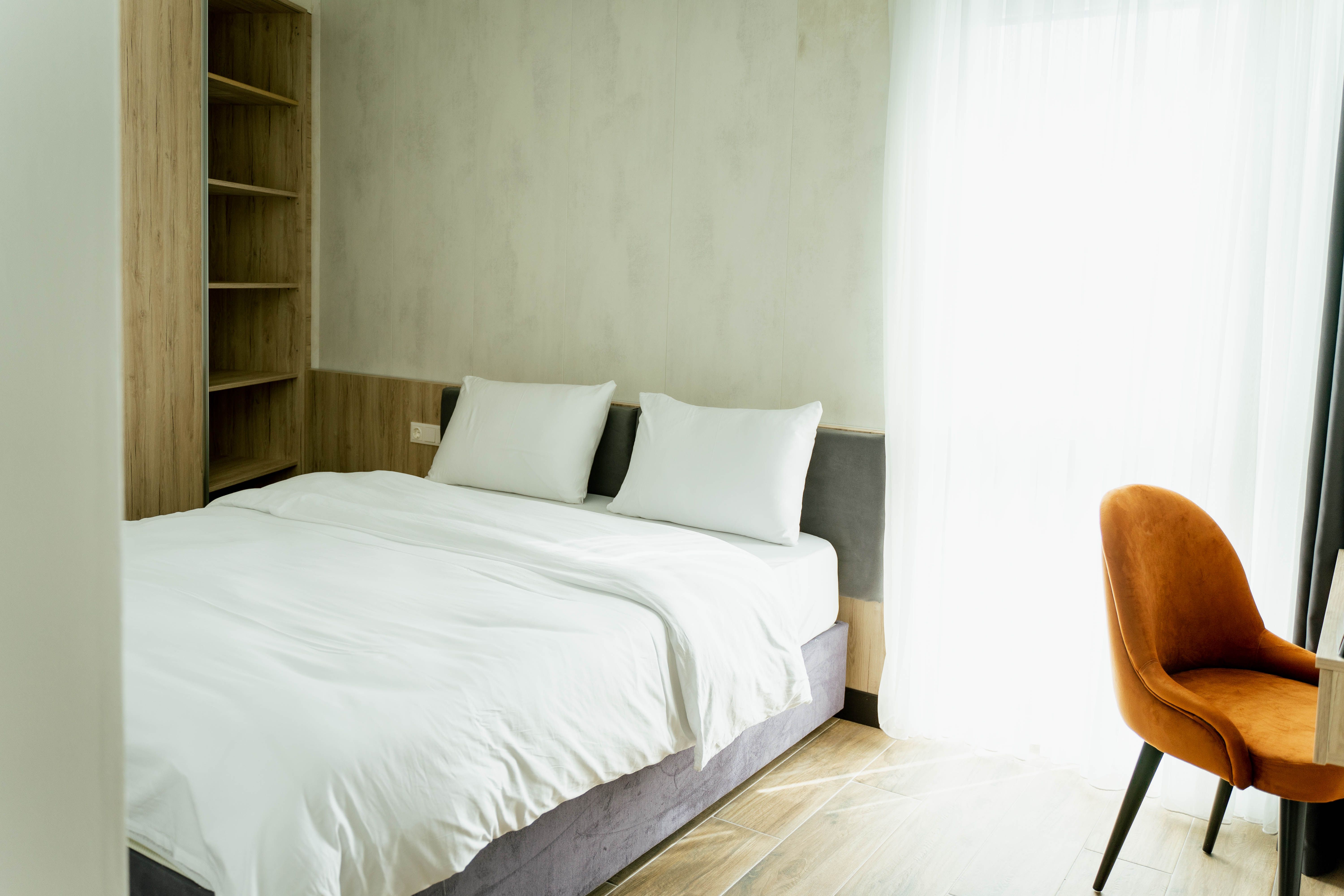 Modern hotel room with double bed, white sheets, wooden shelf and orange chair in front of a window with curtains.