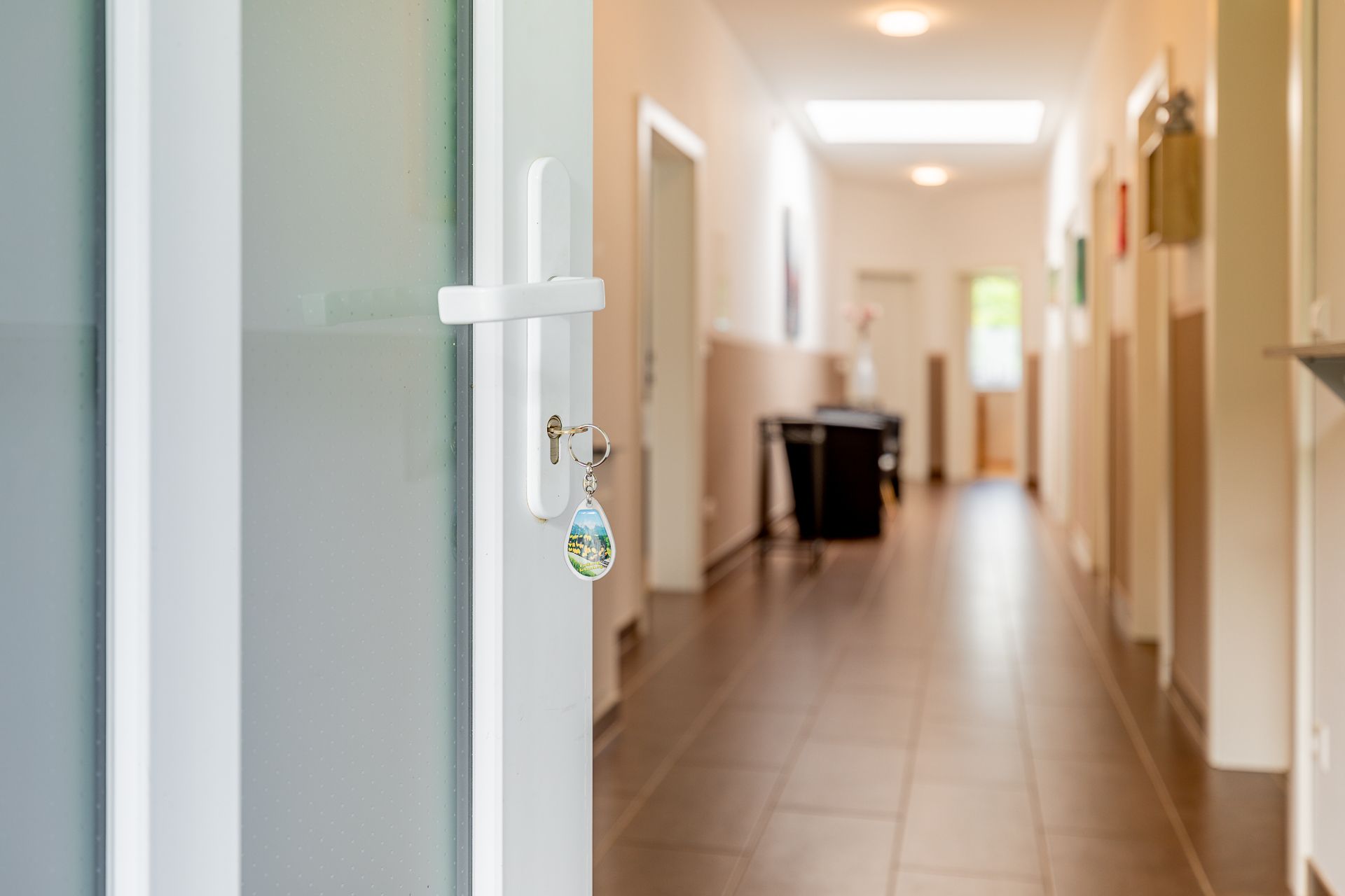 A key is inserted in a white door that leads to a bright hallway with a tiled floor.