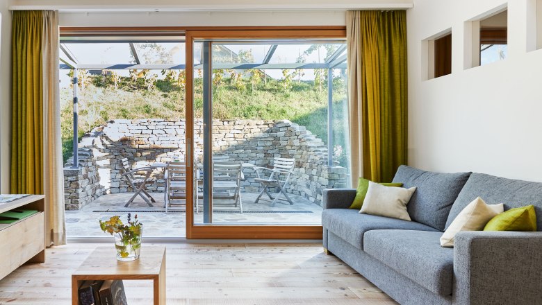 Interior view of a modern living room with gray sofa, wooden floor and large windows leading onto a terrace with stone wall and garden furniture.