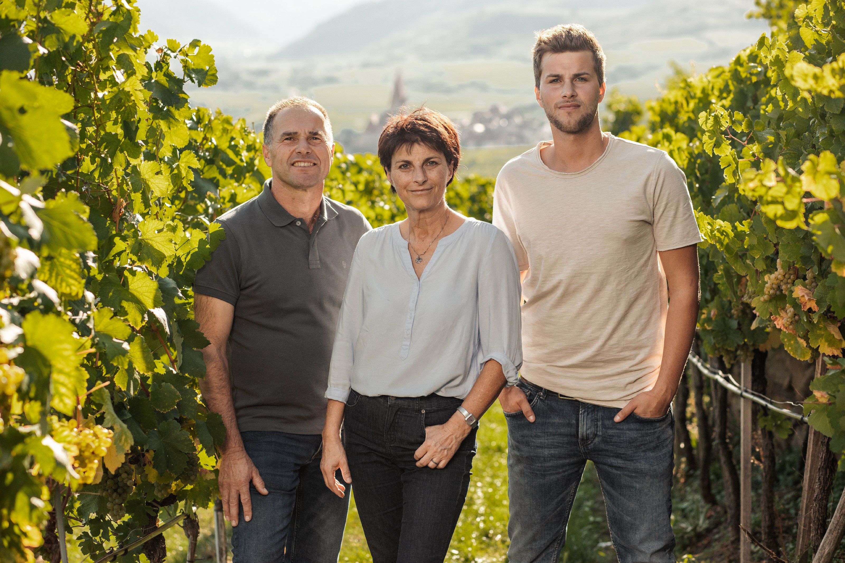Three people are standing in a vineyard, surrounded by green vines, with a village in the background.