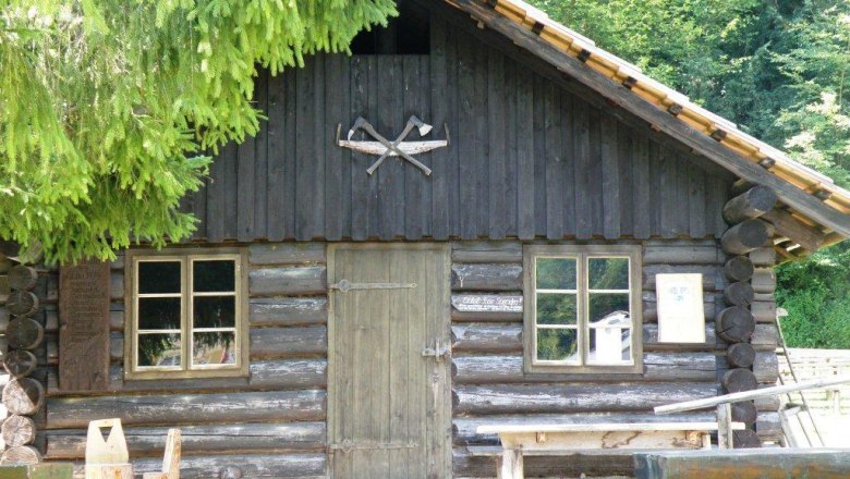 Wooden hut with two windows and a door, surrounded by trees.