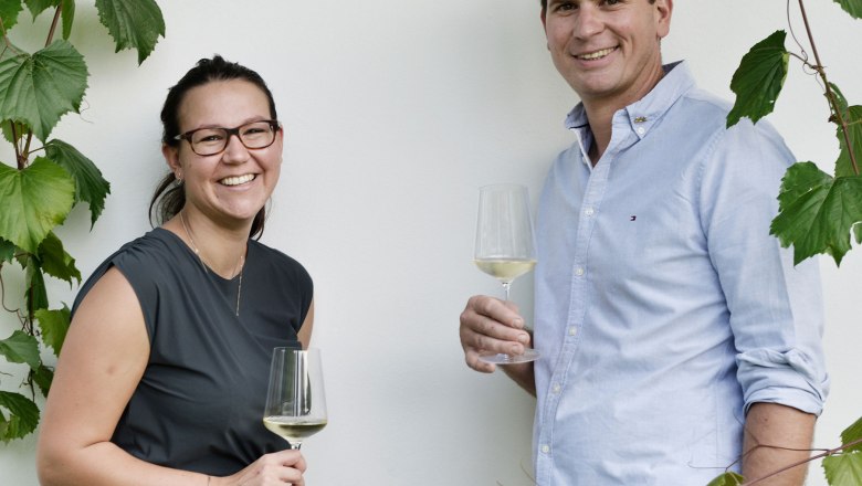 Two people with wine glasses in front of a white wall and grapevines.