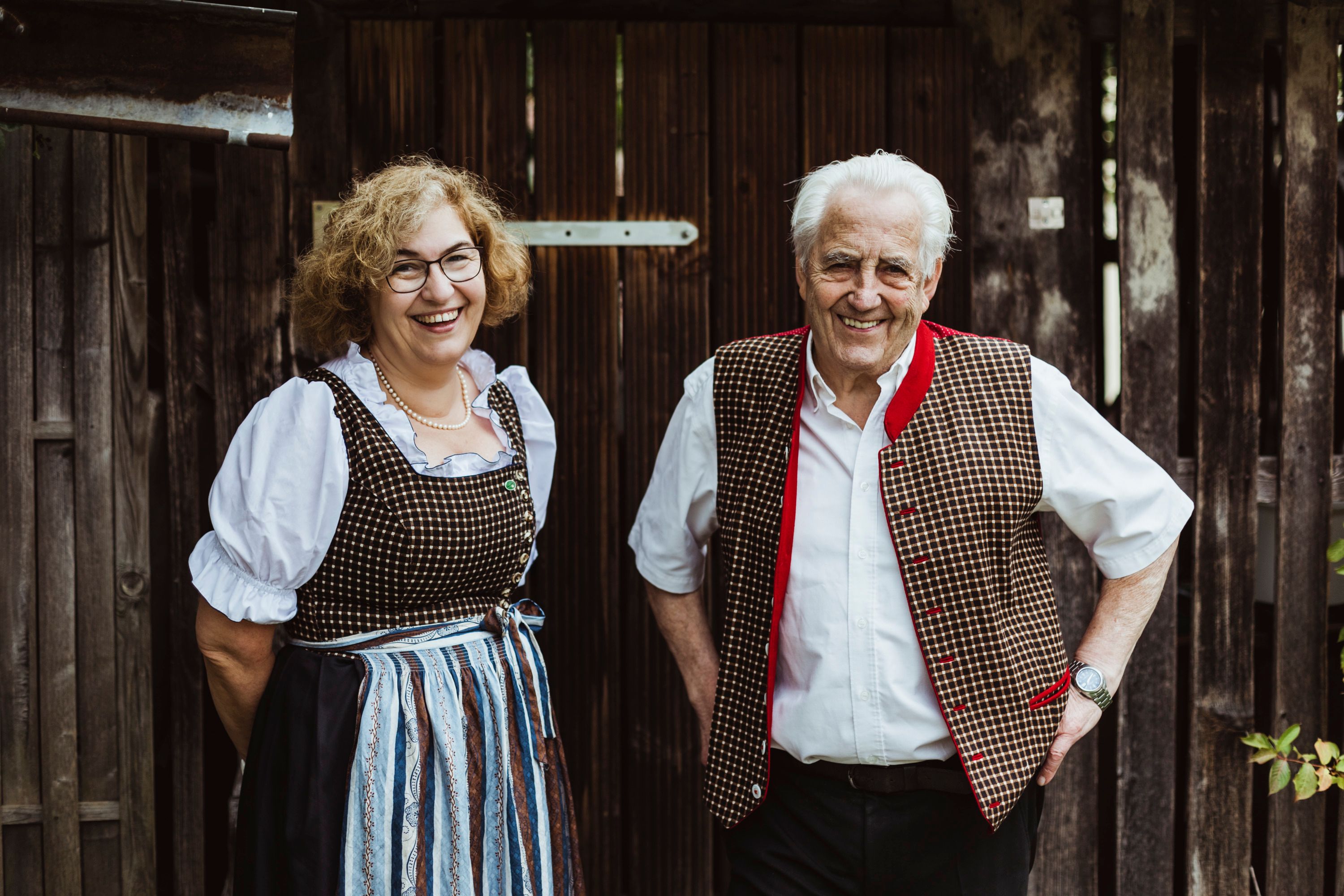 Two people in traditional costume in front of a wooden wall, smiling.