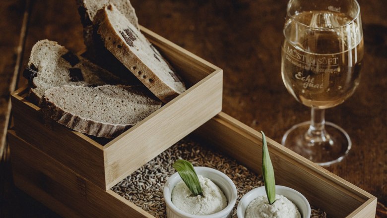A wooden tray with slices of bread, two small bowls of spread and a glass of white wine on a wooden table.