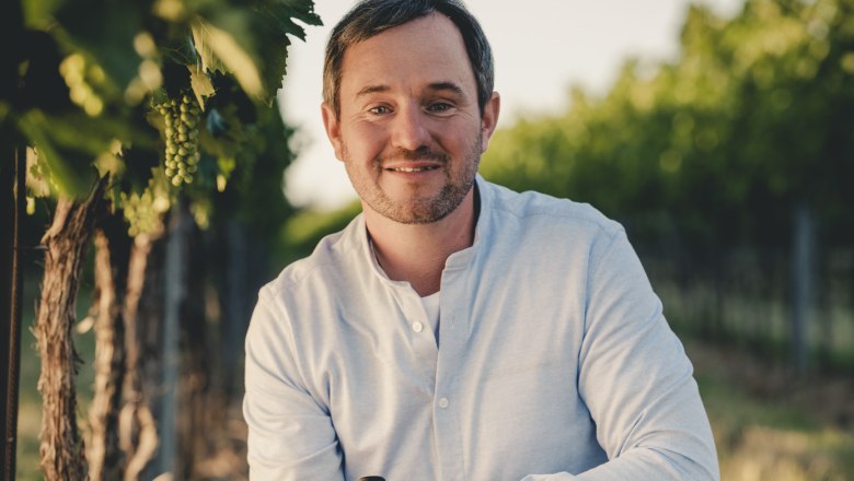A man kneels in a vineyard and holds a bottle of wine in his hand.