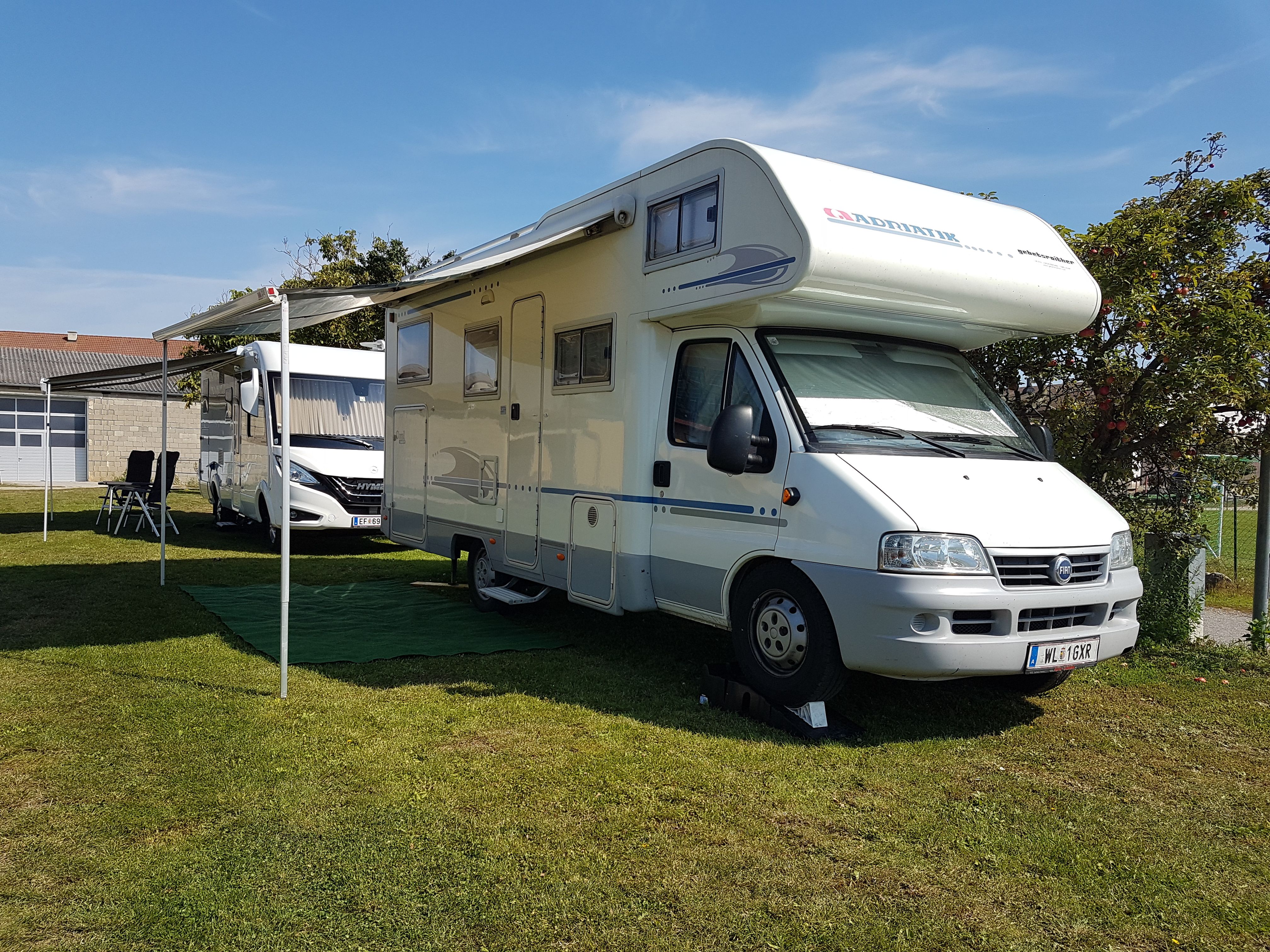 A white motorhome with an extended awning is parked on a meadow next to another vehicle.