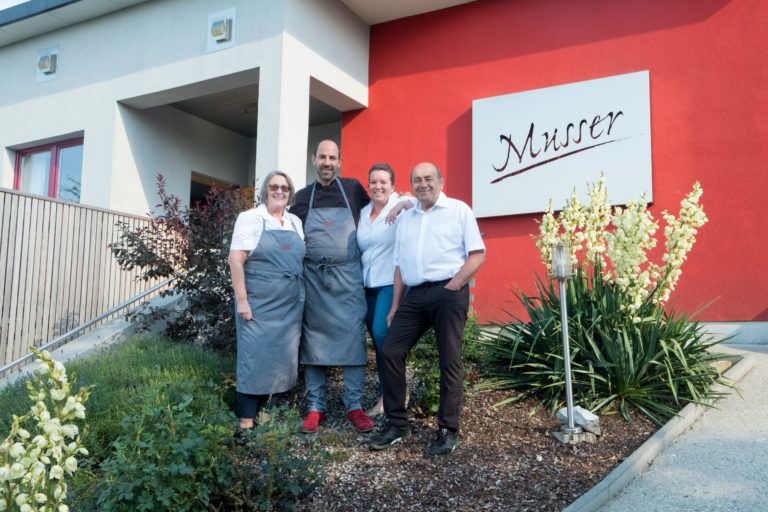 Four people are standing in front of a building with a sign saying 'Musser'.
