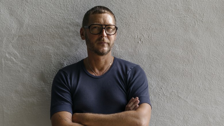 Man with glasses and tattooed arms stands in front of a white wall.