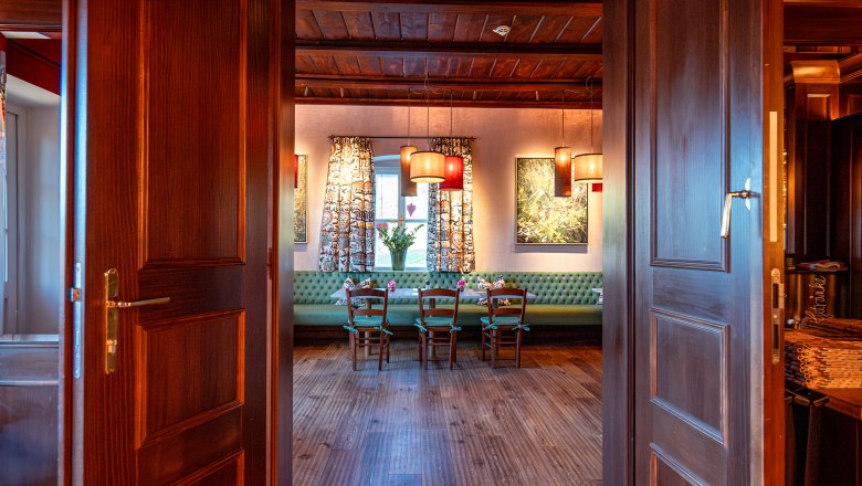 View through open wooden doors into a cozy room with wooden floor, table and chairs, decorated with lamps and curtains.