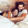 A laughing couple lie together in bed in front of a wooden wall.