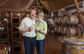 A couple is standing in a distillery with wooden barrels and holding glasses of spirits.
