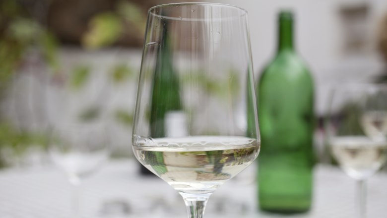 A wine glass with white wine stands on a table, a green wine bottle in the background.