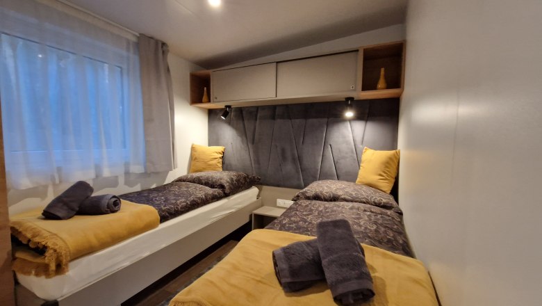 Two single beds in a cozy chalet room with yellow blankets and pillows, grey towels and a window with a curtain.