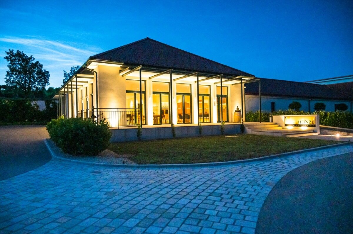 A modern building with glass fronts, illuminated at dusk, surrounded by a paved path and green spaces.