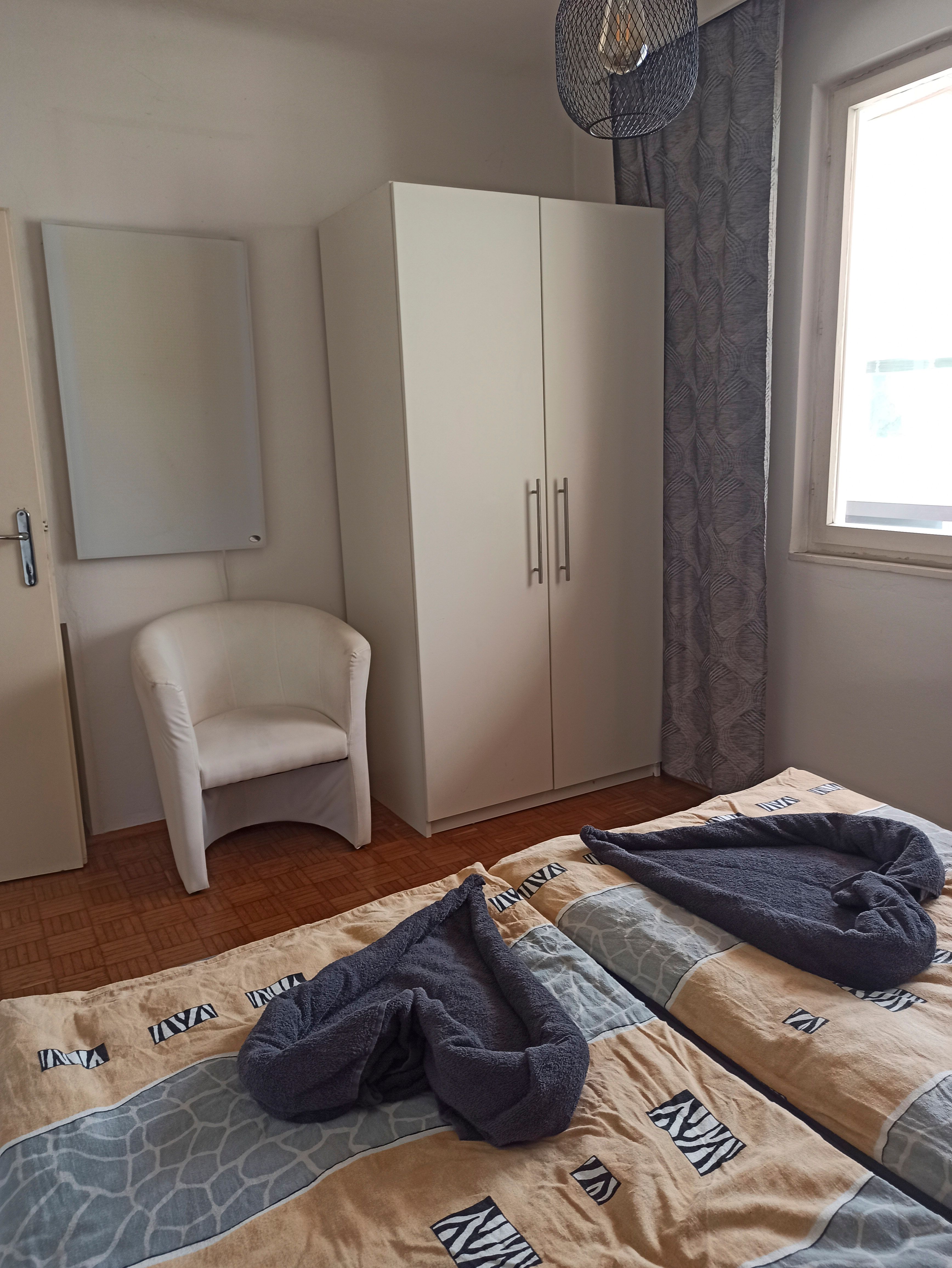 A guest room with two beds, a white wardrobe, an armchair and a window with a curtain.