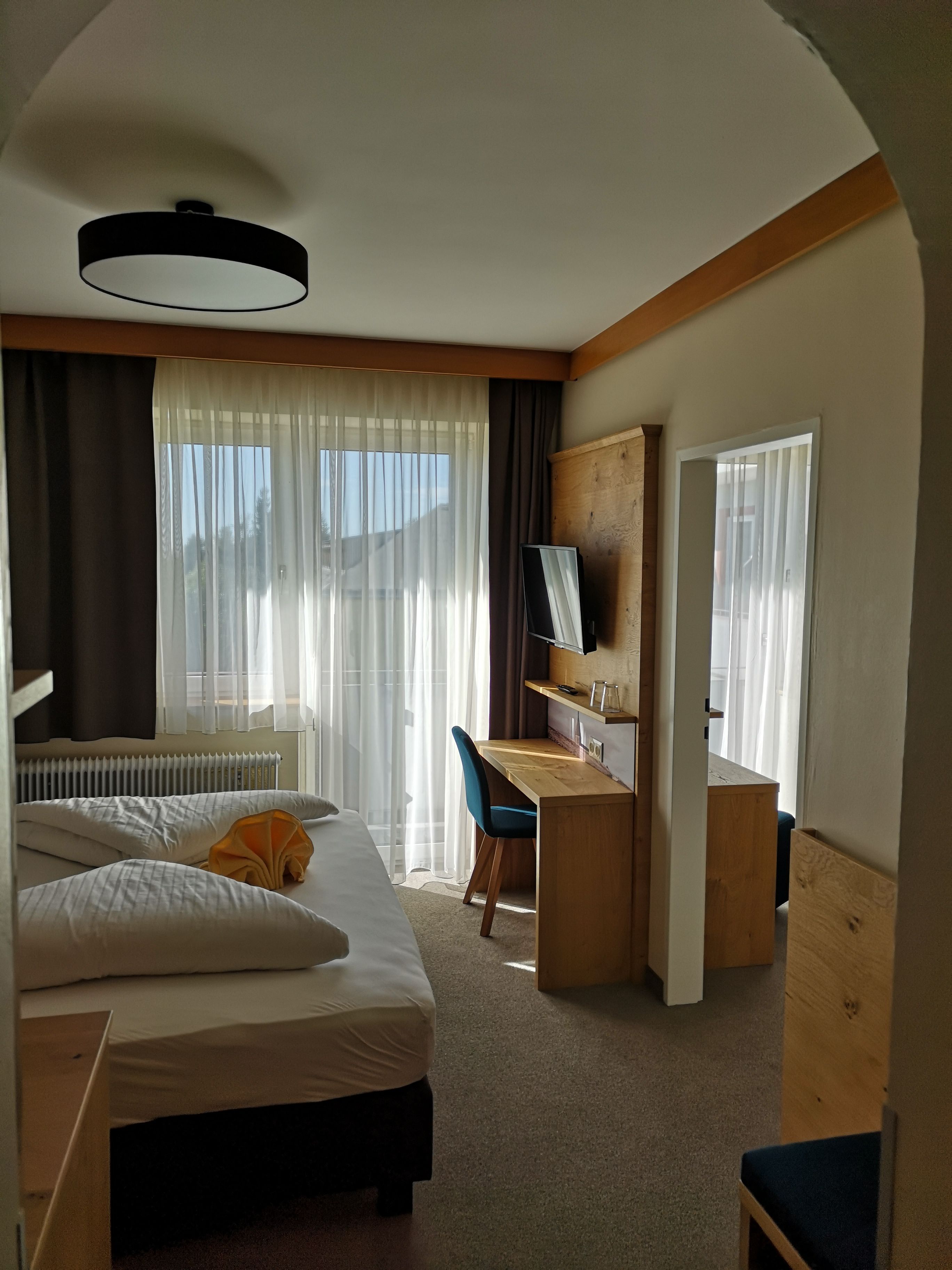 A modern double room with a bed, desk, chair and TV. Large windows with curtains let in daylight.