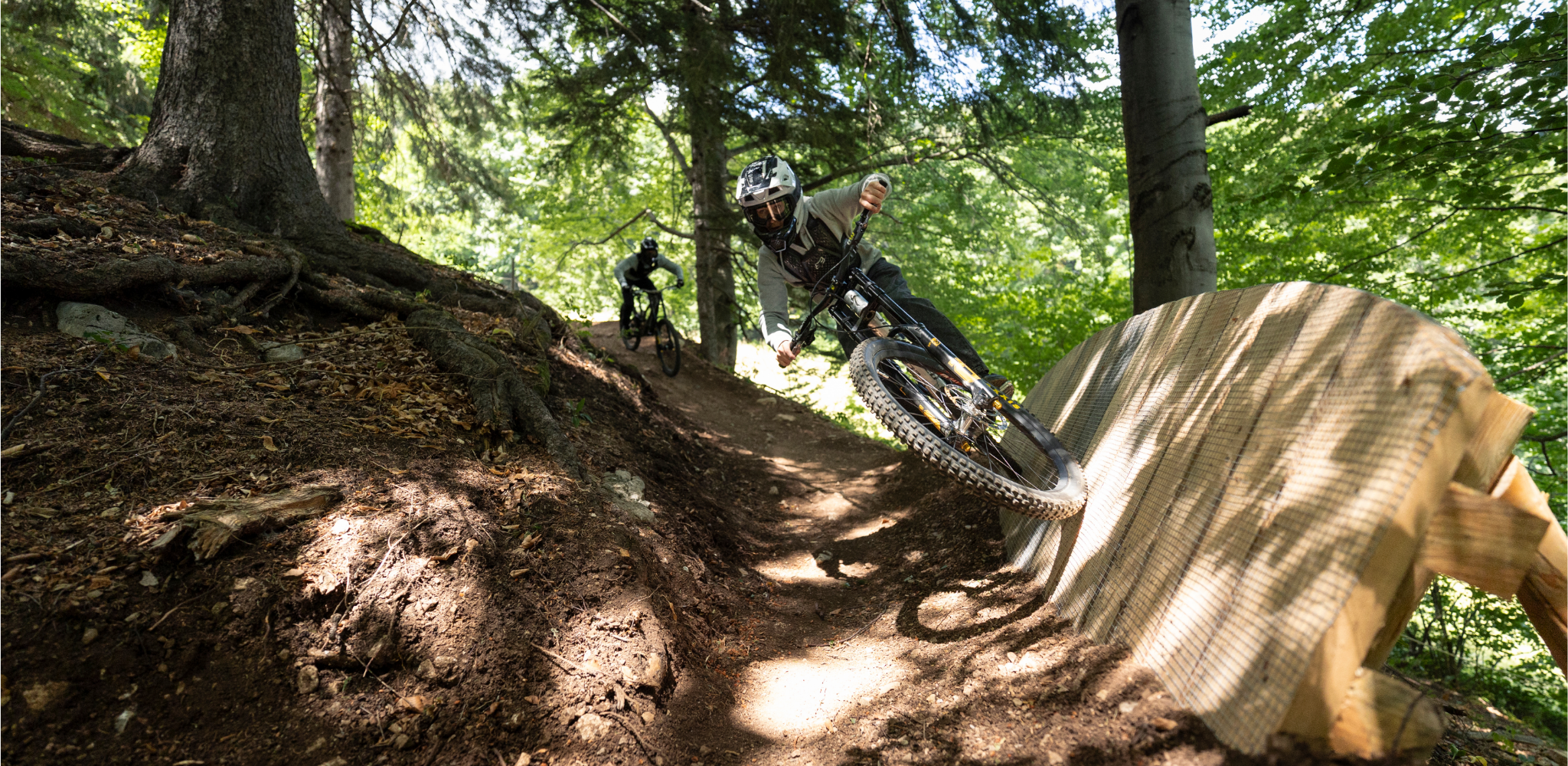 Two mountain bikers are riding on a forest trail with a wooden ramp.