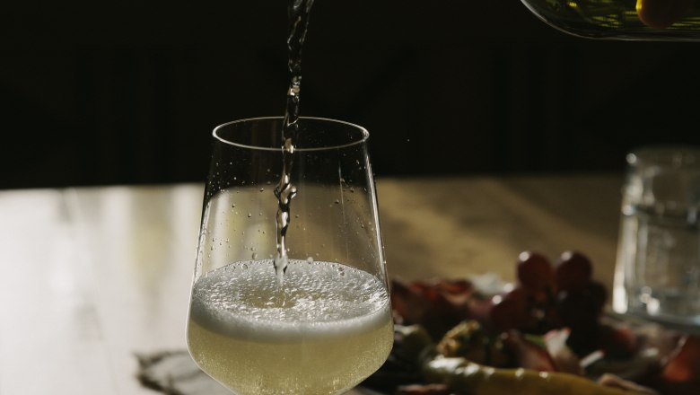 A glass of white wine is poured at a table with a plate full of snacks.