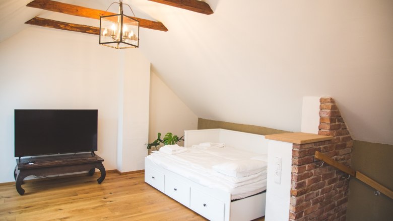 An attic room with a wooden floor, a white bed, a TV on a table and exposed wooden beams on the ceiling.