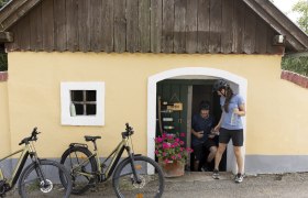 Two people with bicycles in front of a small yellow building with a wooden roof and flowers.