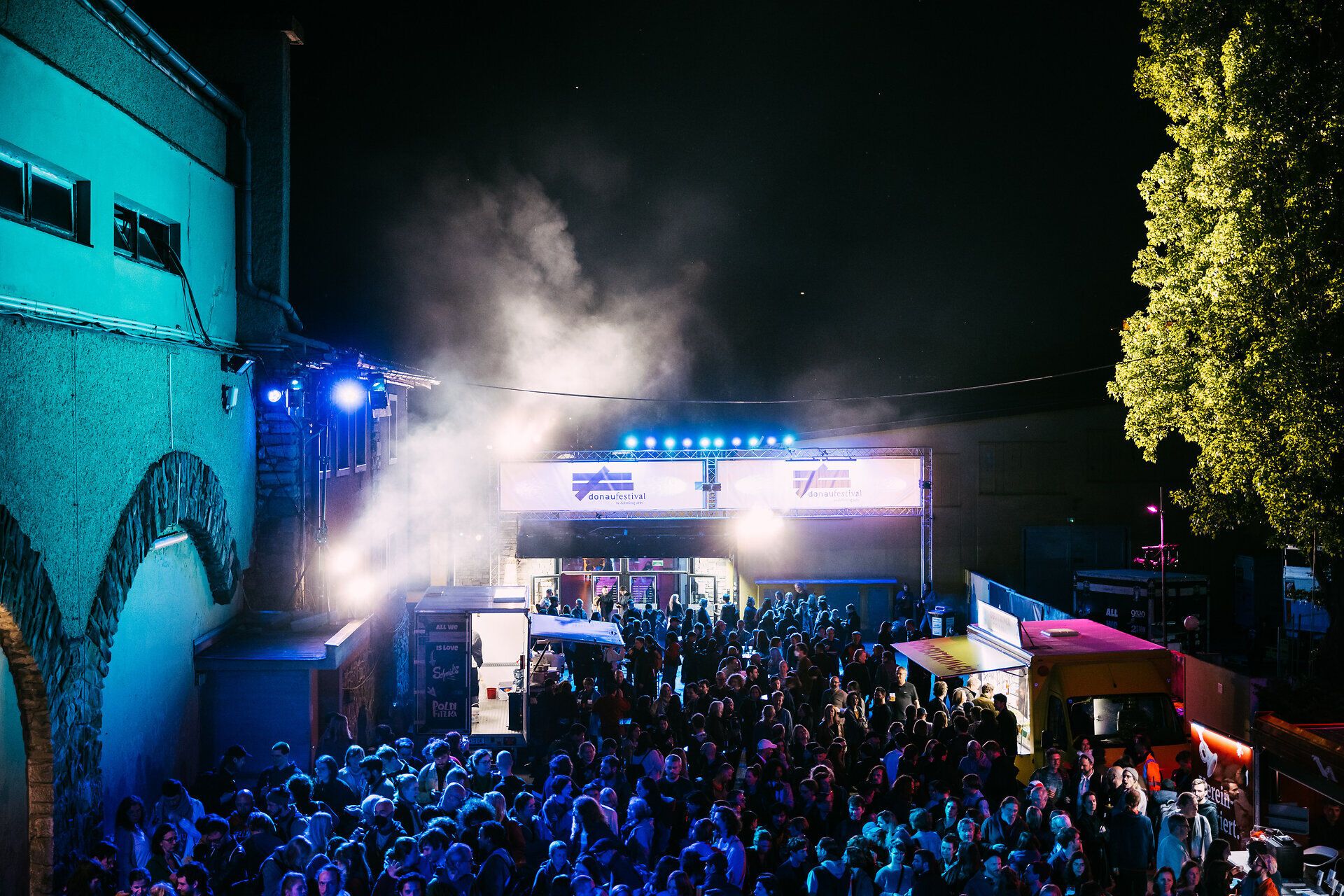 Nächtliche Festivalatmosphäre am Messegelände des donaufestivals: dicht gedrängtes Publikum vor Bühnen und Foodtrucks, beleuchtet von blauem Licht, Nebel und Scheinwerfern.