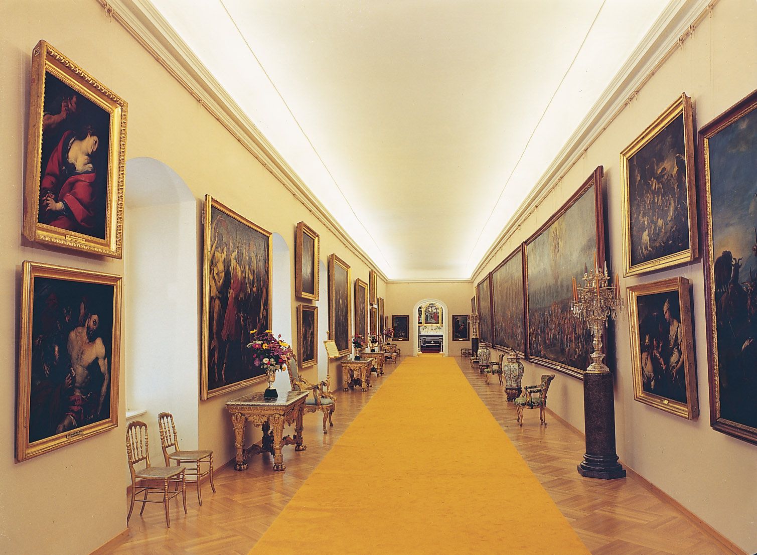 Long, elegant gallery space with paintings on the walls and a yellow carpet in the middle.