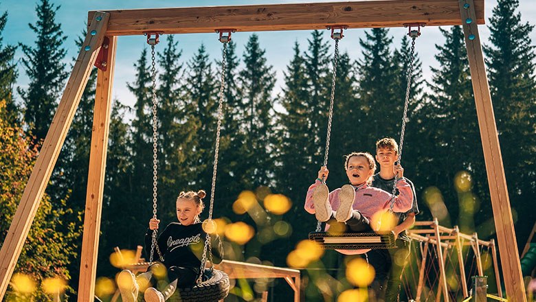 Three children swinging on a large outdoor swing surrounded by trees.