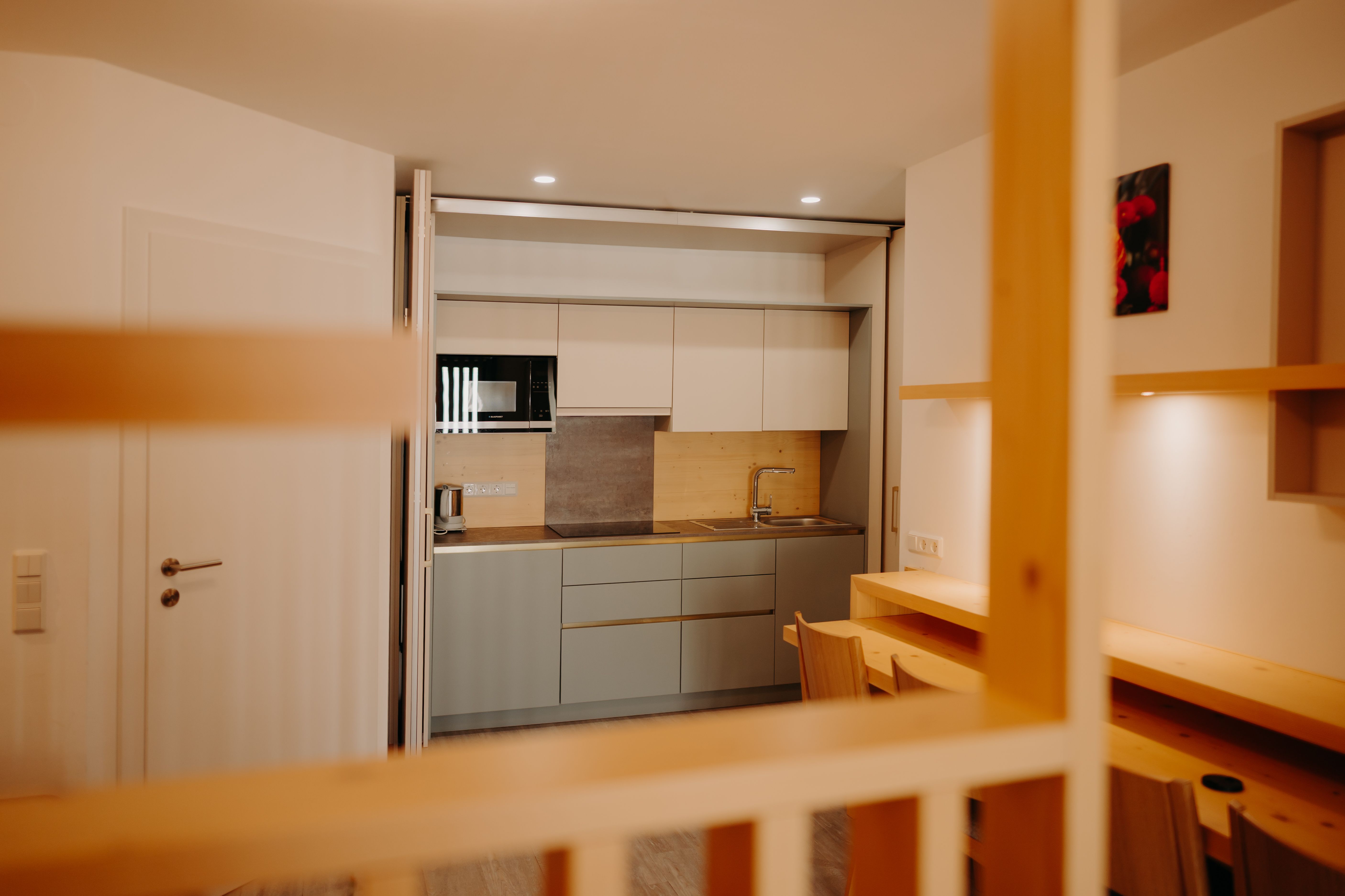A modern kitchenette in an apartment, decorated in light colors. It features a sink, a microwave, a large stovetop, and an electric kettle. The kitchen can be hidden behind a sliding partition. In front of the kitchen, there is a table against the wall and four armchairs.