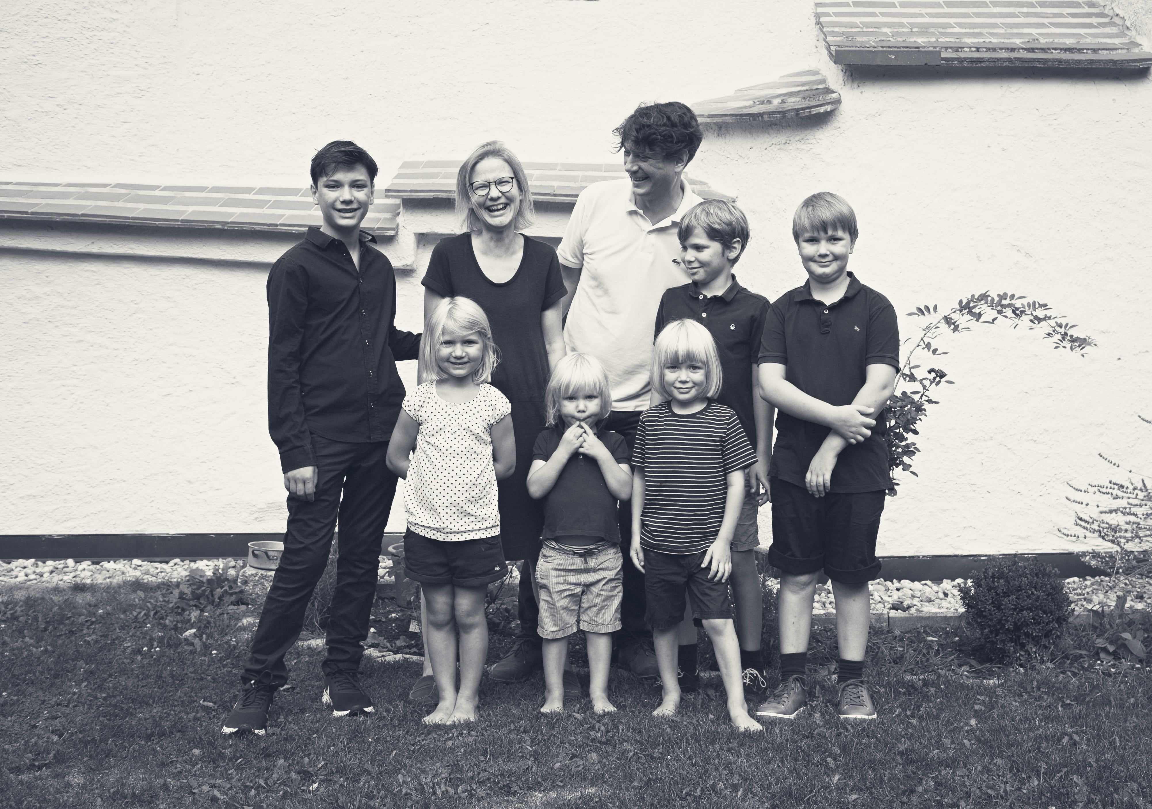 A family of seven stands smiling in front of a white wall outside.
