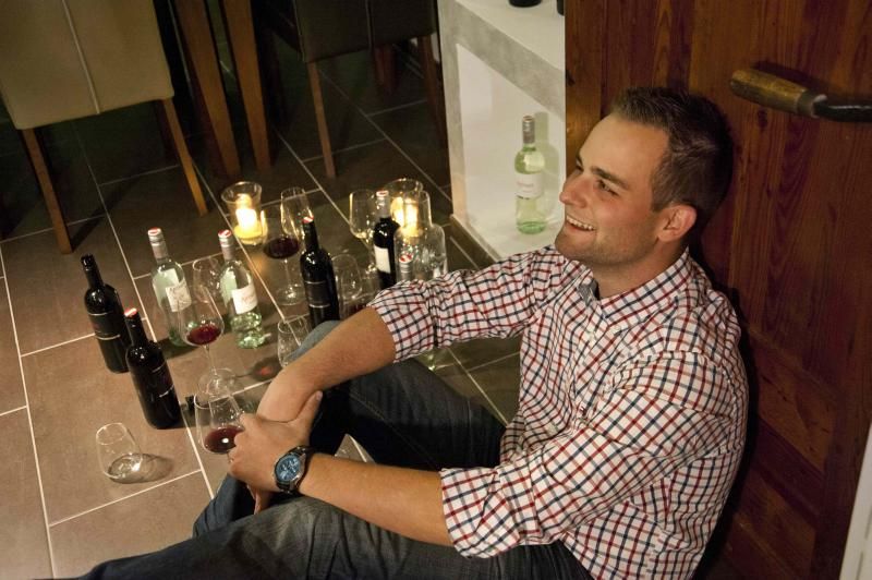 A man sits smiling on the floor, surrounded by bottles of wine and glasses.