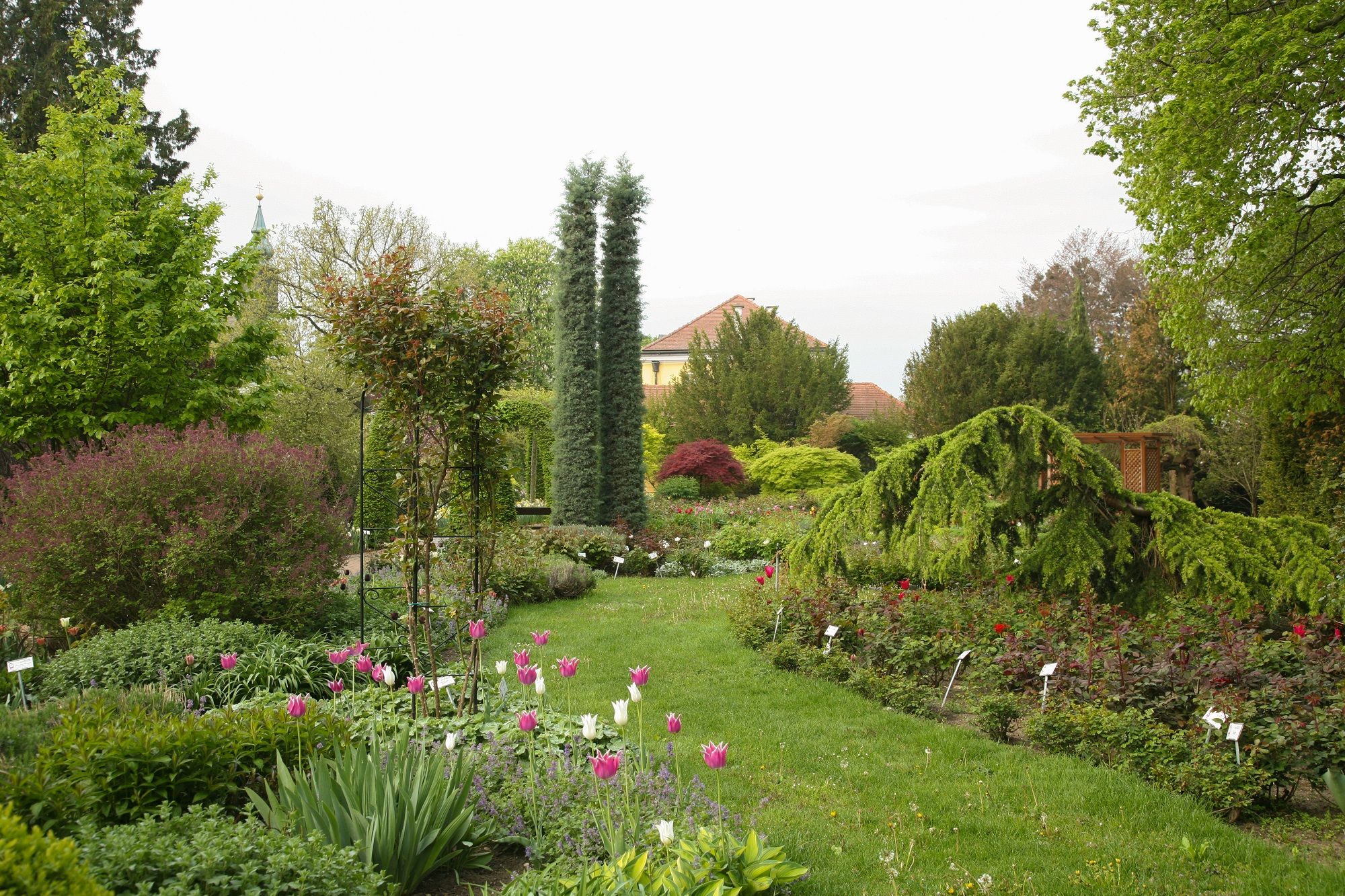A well-tended garden with colorful flowers, trees and shrubs, in the background a building with a red roof.