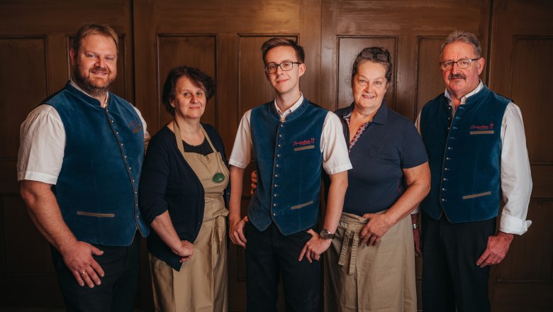 Five people in traditional dress stand in front of a wooden wall.