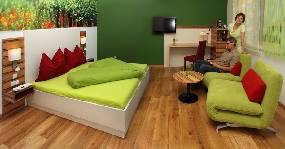 A modern guest room with a green bed, sofa and wooden floor. There are two people in the room, one sitting on the sofa, the other standing next to it.