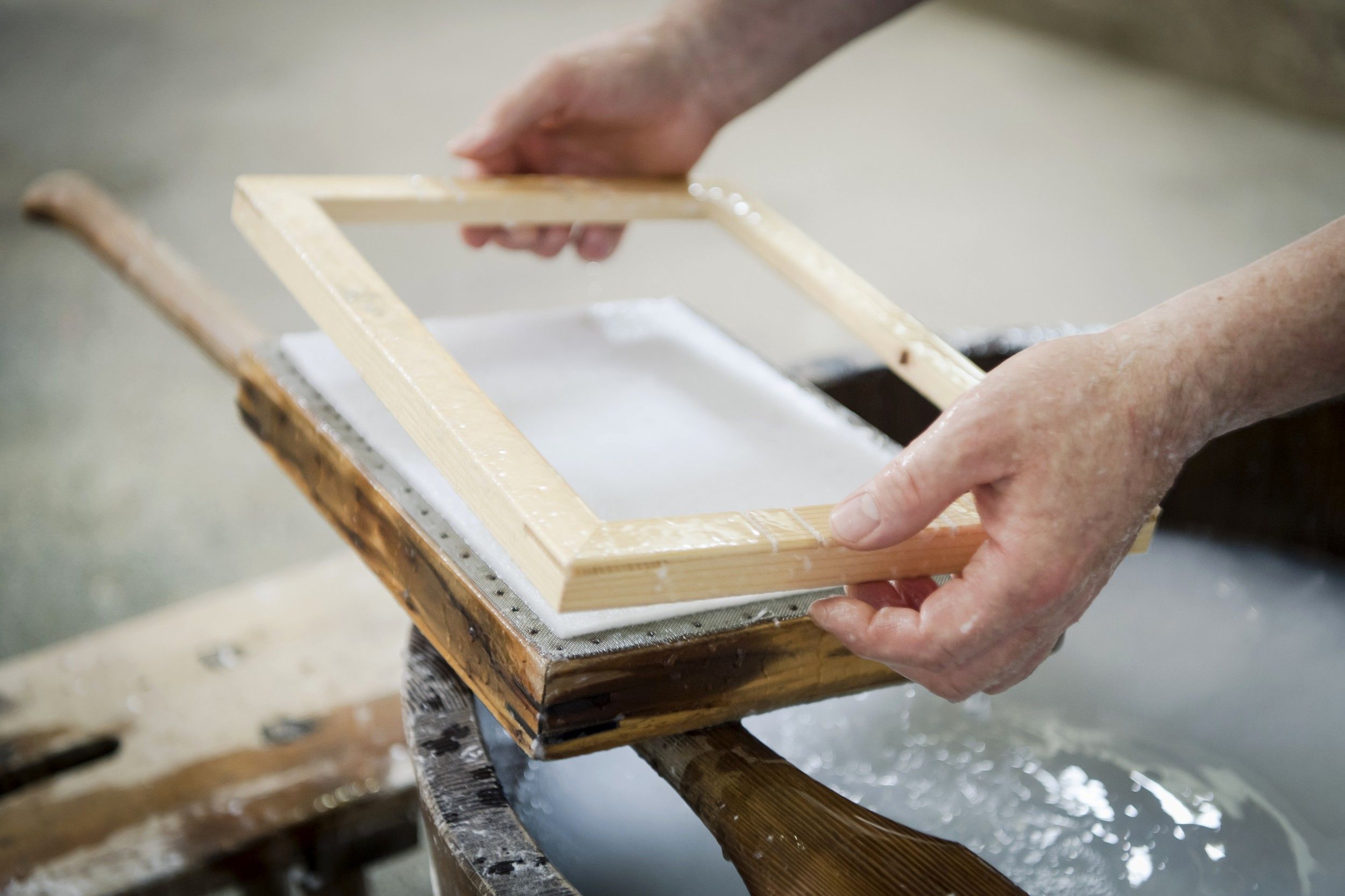 Close-up of hands holding a wooden frame over a paper pulp in a traditional paper mill.