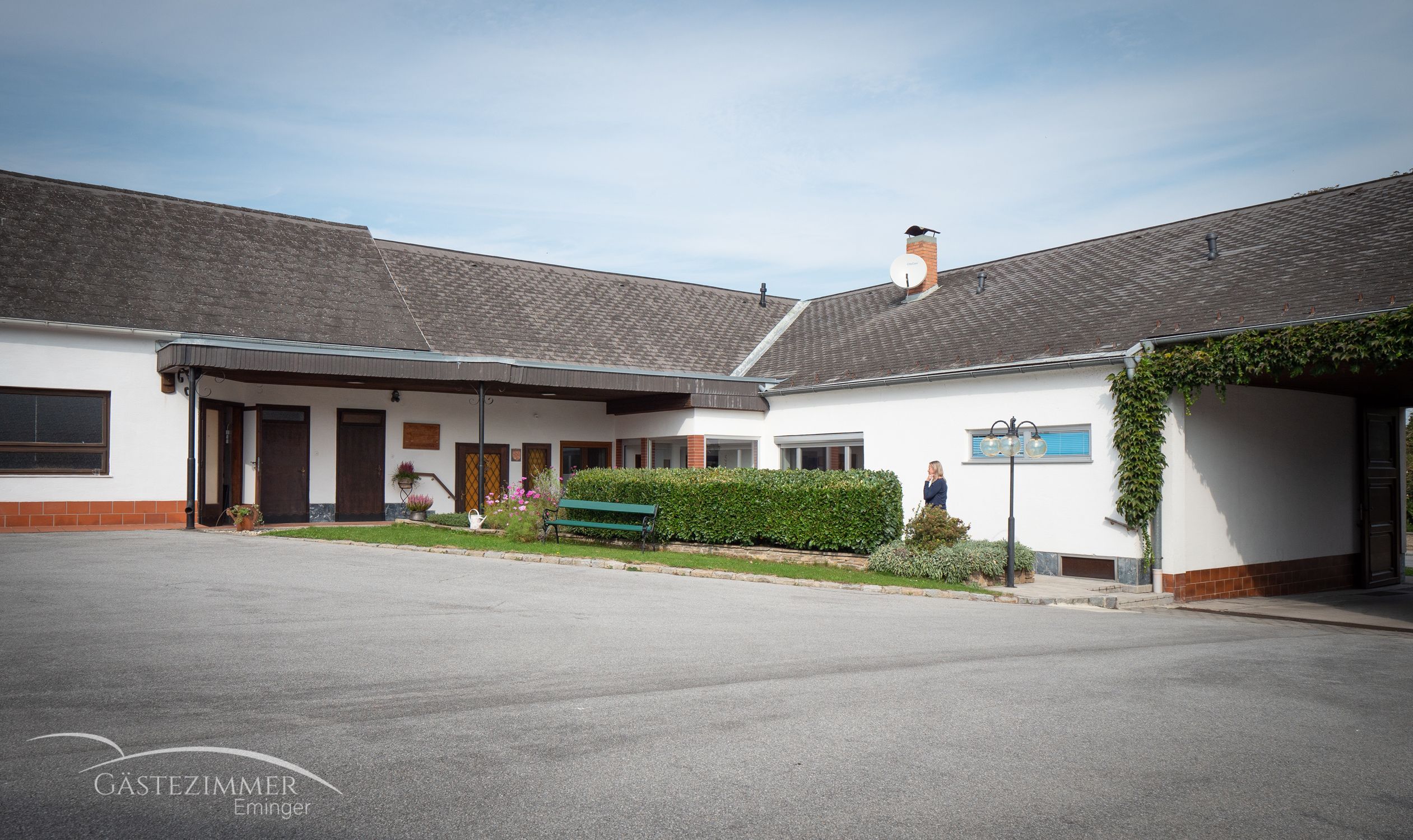 A courtyard with a building, parking spaces and a hedge. A person is standing next to the hedge. A satellite dish can be seen on the building.
