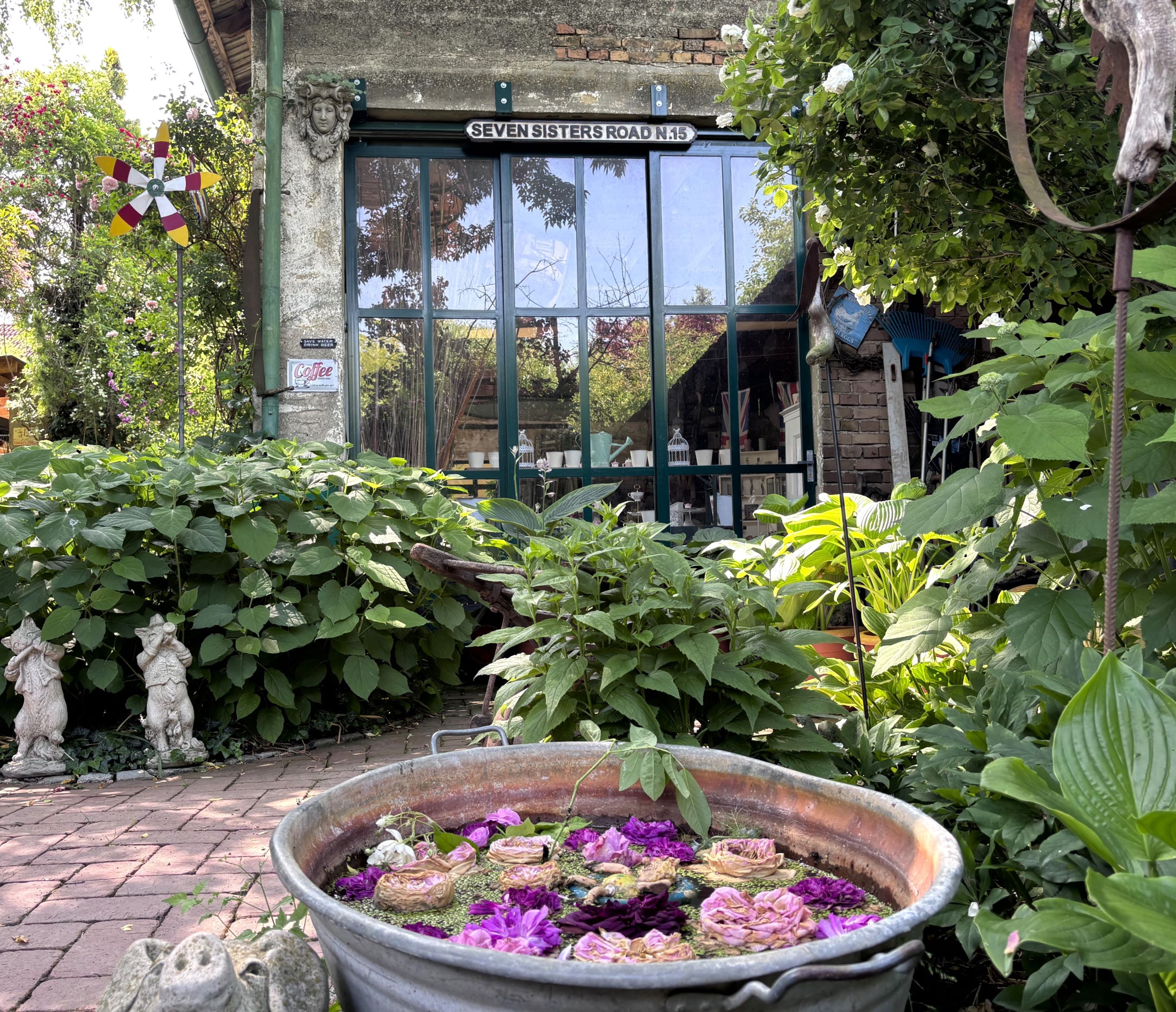 A cottage garden with a zinc container full of flowers, surrounded by lush greenery and a glass front with the inscription 'Seven Sisters Road N15'.