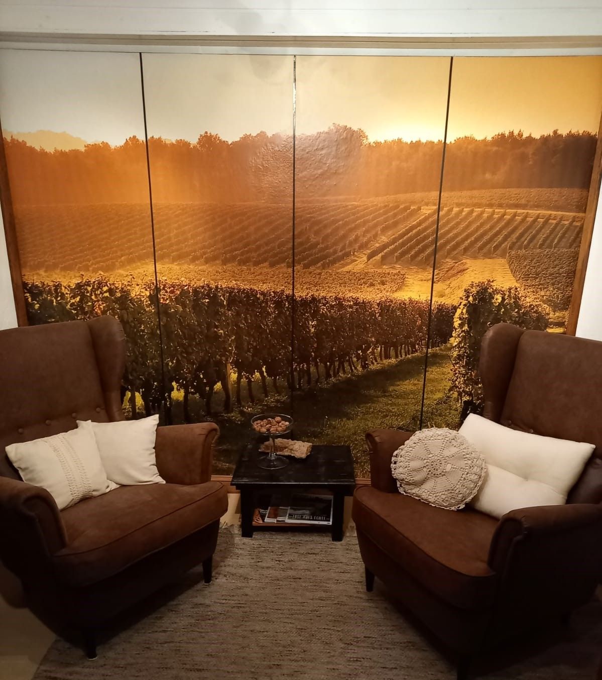 Two brown armchairs with cushions in front of a wall with a landscape photo wallpaper of a vineyard.