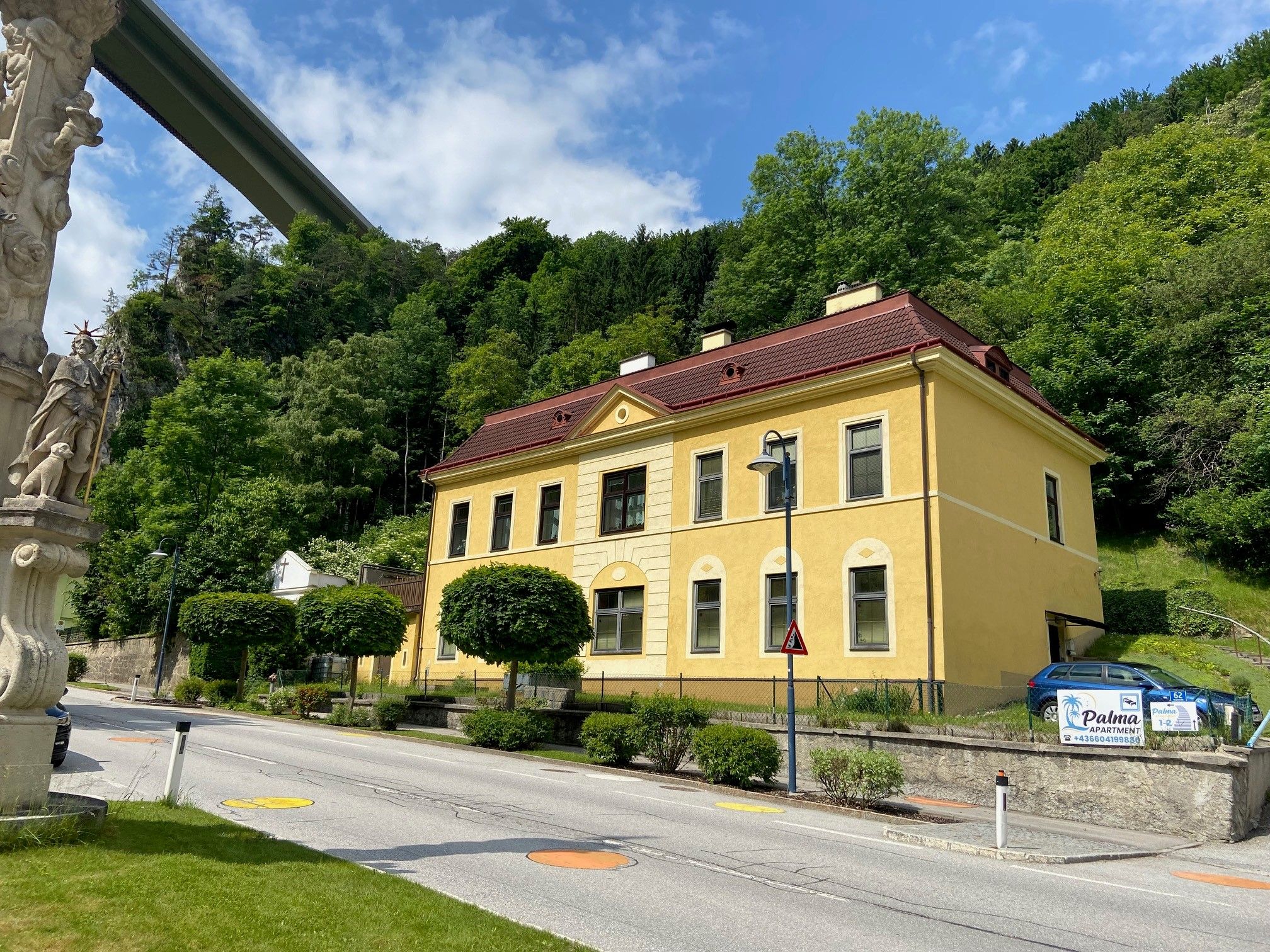Yellow building with a red roof next to a road, surrounded by trees and a statue in the foreground.