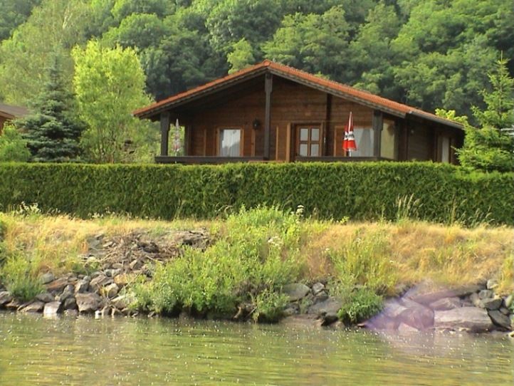 A wooden holiday home with a red roof, surrounded by trees and bushes, stands on the banks of a body of water.
