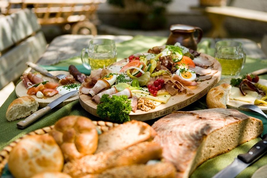 A richly laid table with a selection of cold meats, cheese, bread and beer mugs outside.