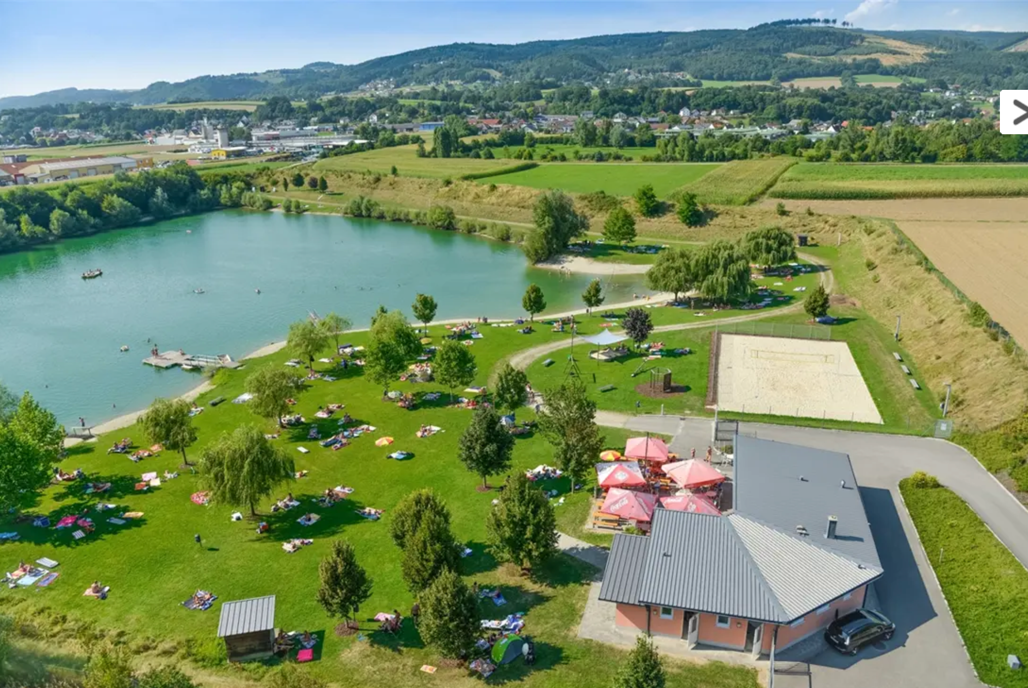 Aerial view of a bathing pond with sunbathing lawn, trees and a building with sunshades.