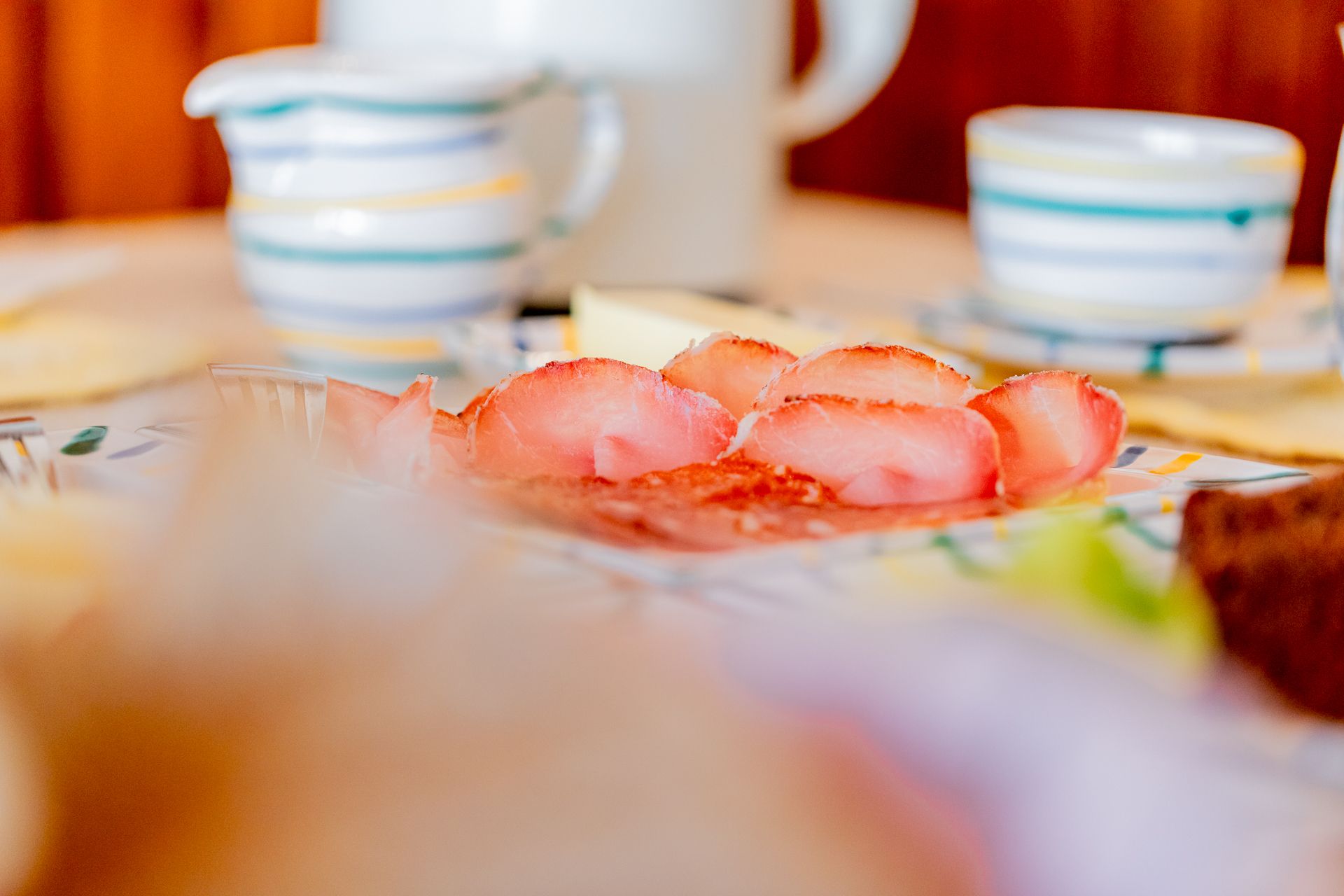 Close-up of a breakfast table with ham, crockery and cups.