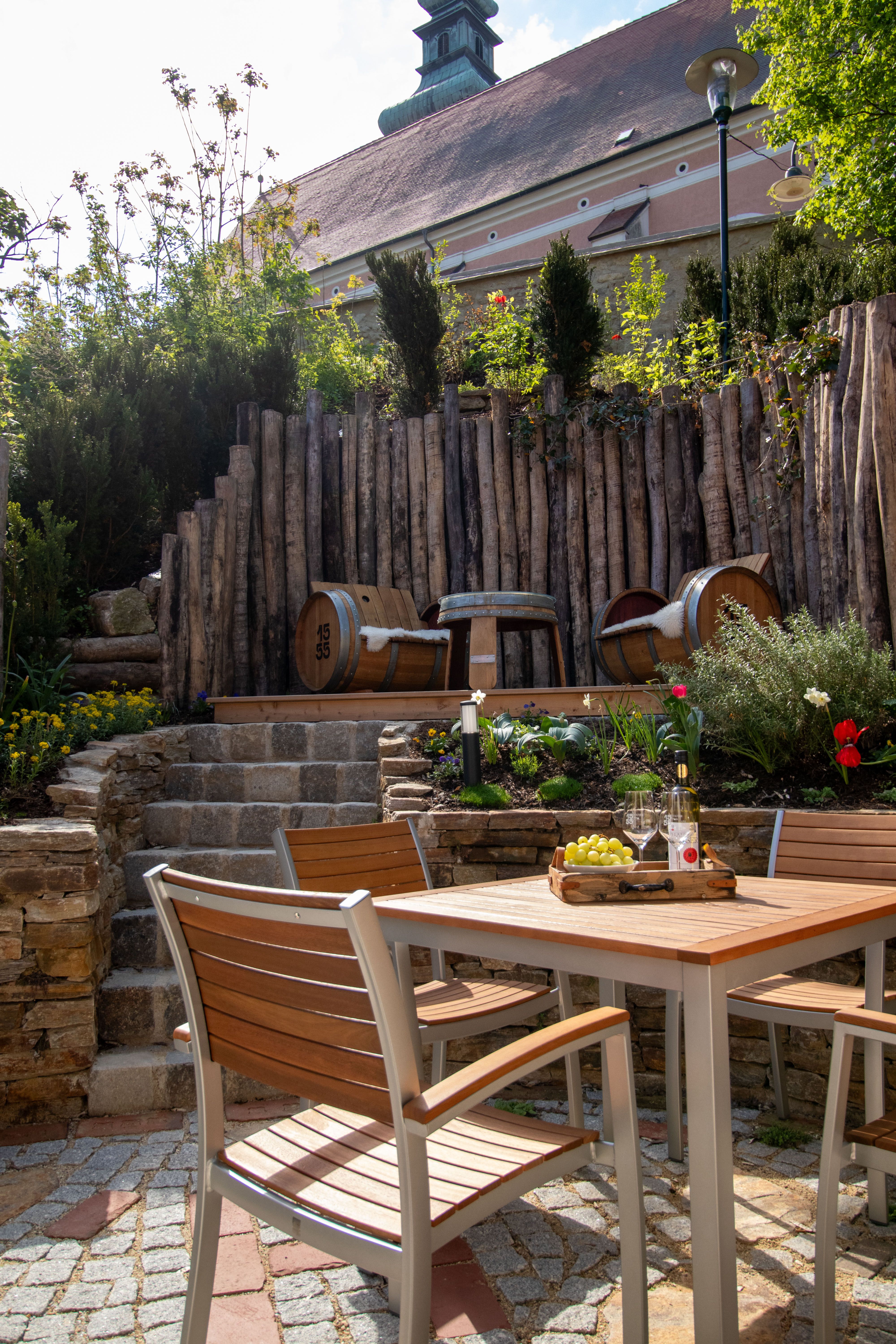 A cozy garden area with wooden table and chairs, wine bottles and glasses. A wooden wall in the background.