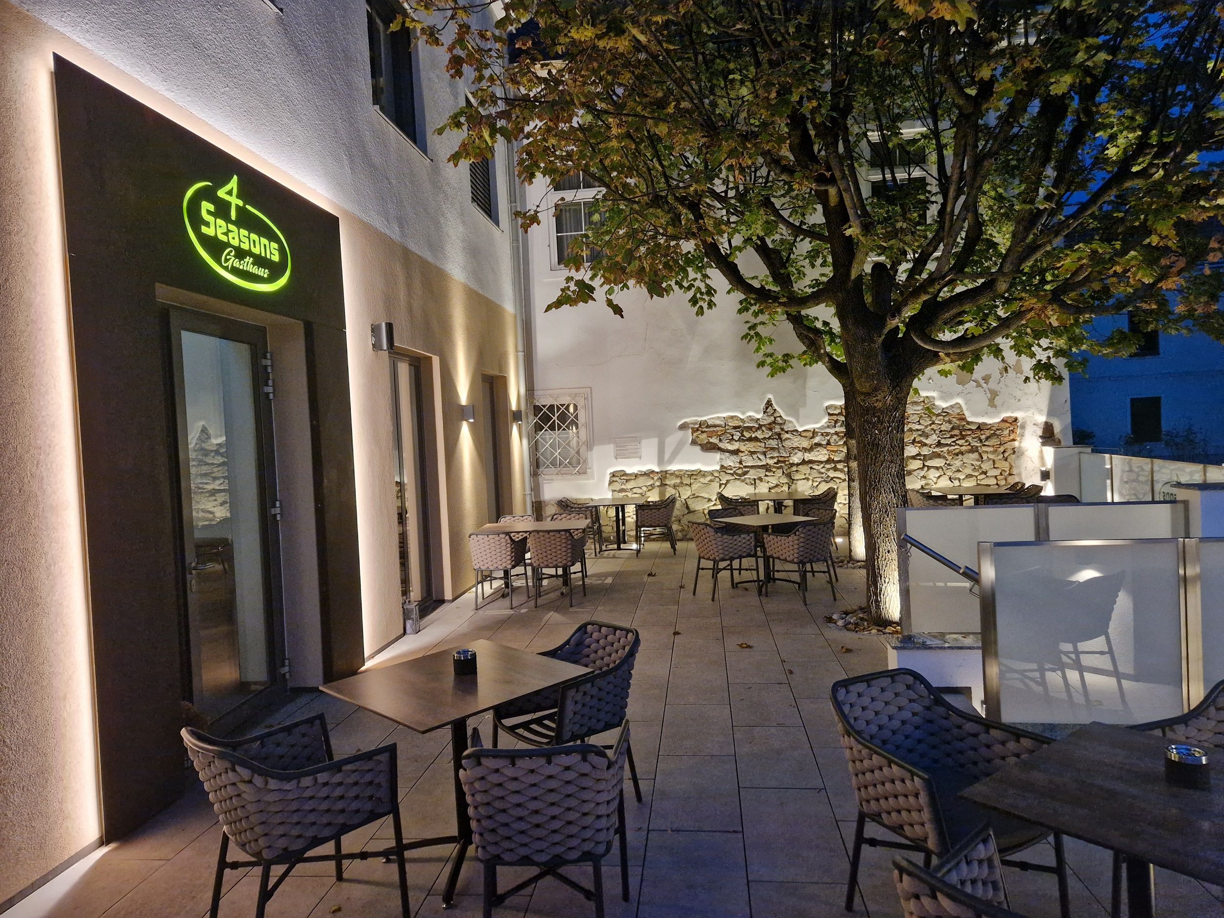 Outdoor terrace of a restaurant with tables and chairs, illuminated at dusk, tree in the background.