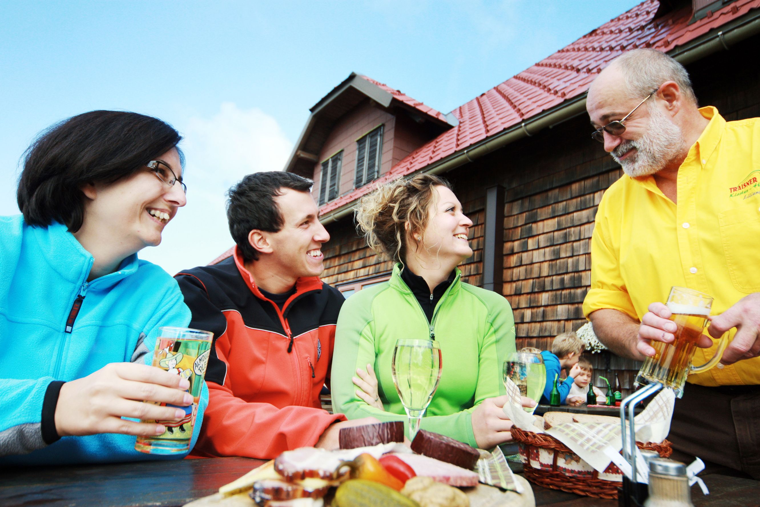 Three people sit laughing at a table in front of a hut while a man in a yellow shirt serves drinks.