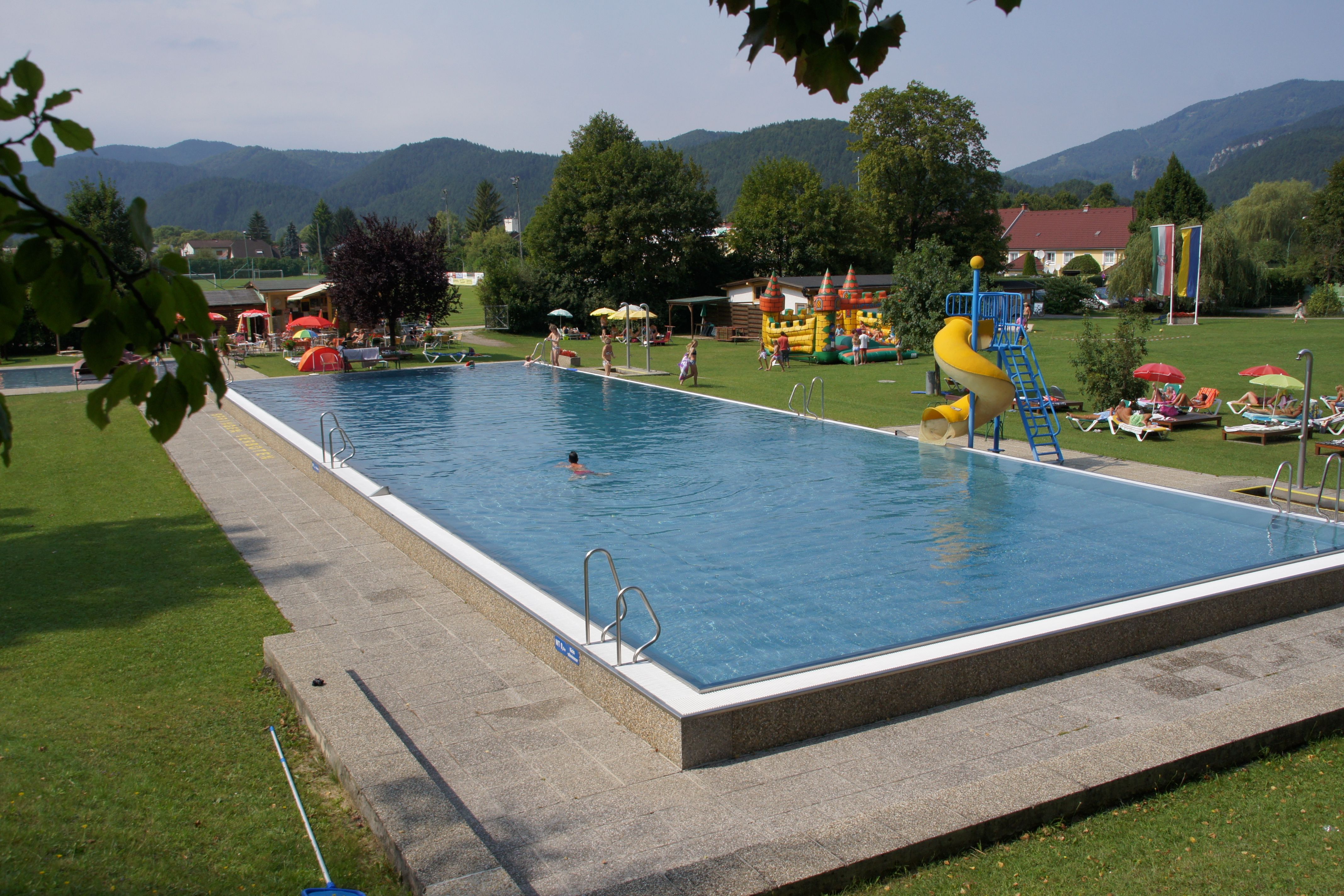 An outdoor pool with swimming pool, slide and sunbathing lawn, surrounded by trees and mountains.