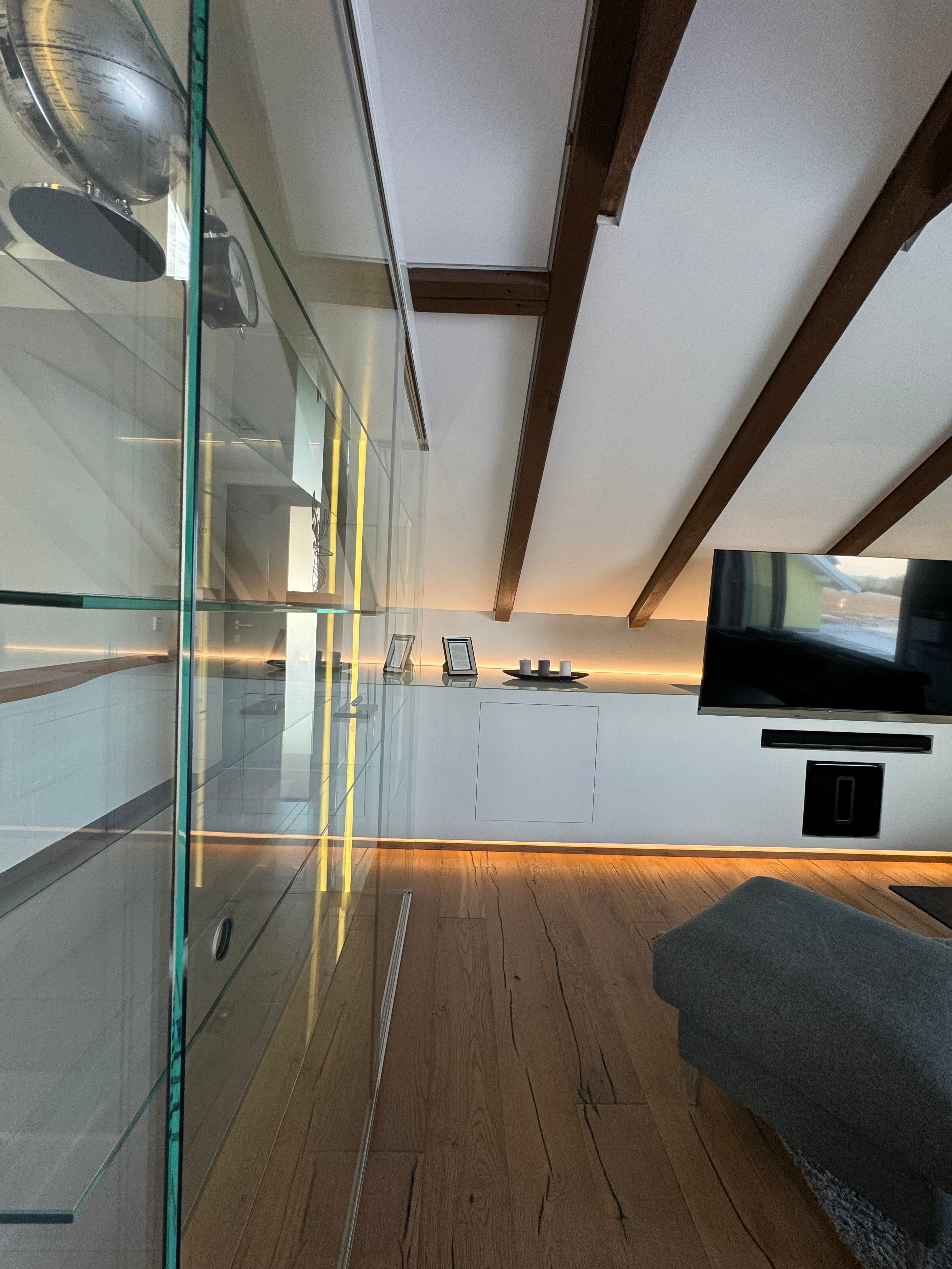 Modern living room with wooden floor, glass cabinet, TV and sloping ceiling with wooden beams.