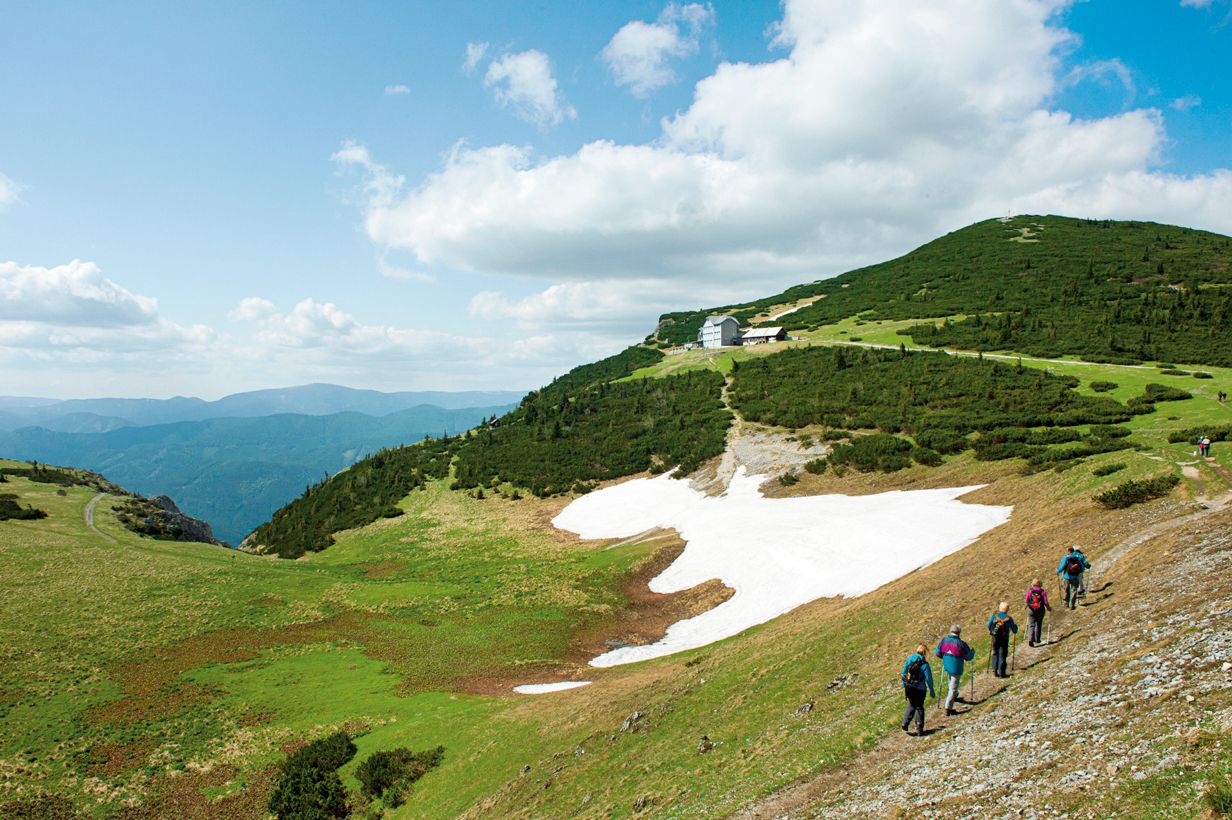 Hikers on the Raxalpe with snow and mountain hut in the background.